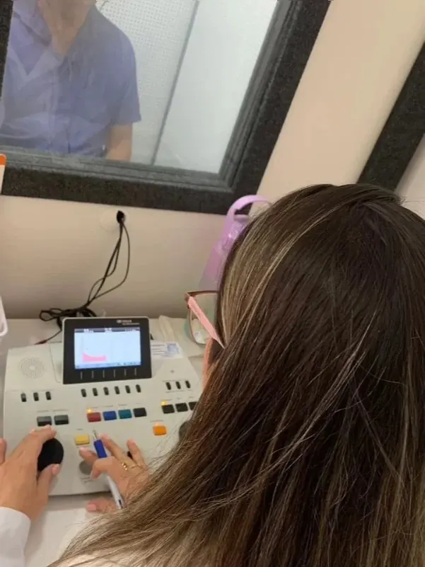 Audiometria sendo realizada na Protec, com a cliente dentro da cabine acústica e a fonoaudióloga operando o audiômetro